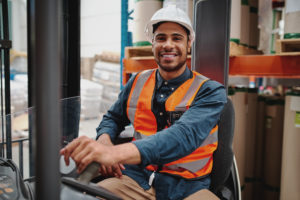 Smiling driver sitting in forklift machine transporting goods in warehouse