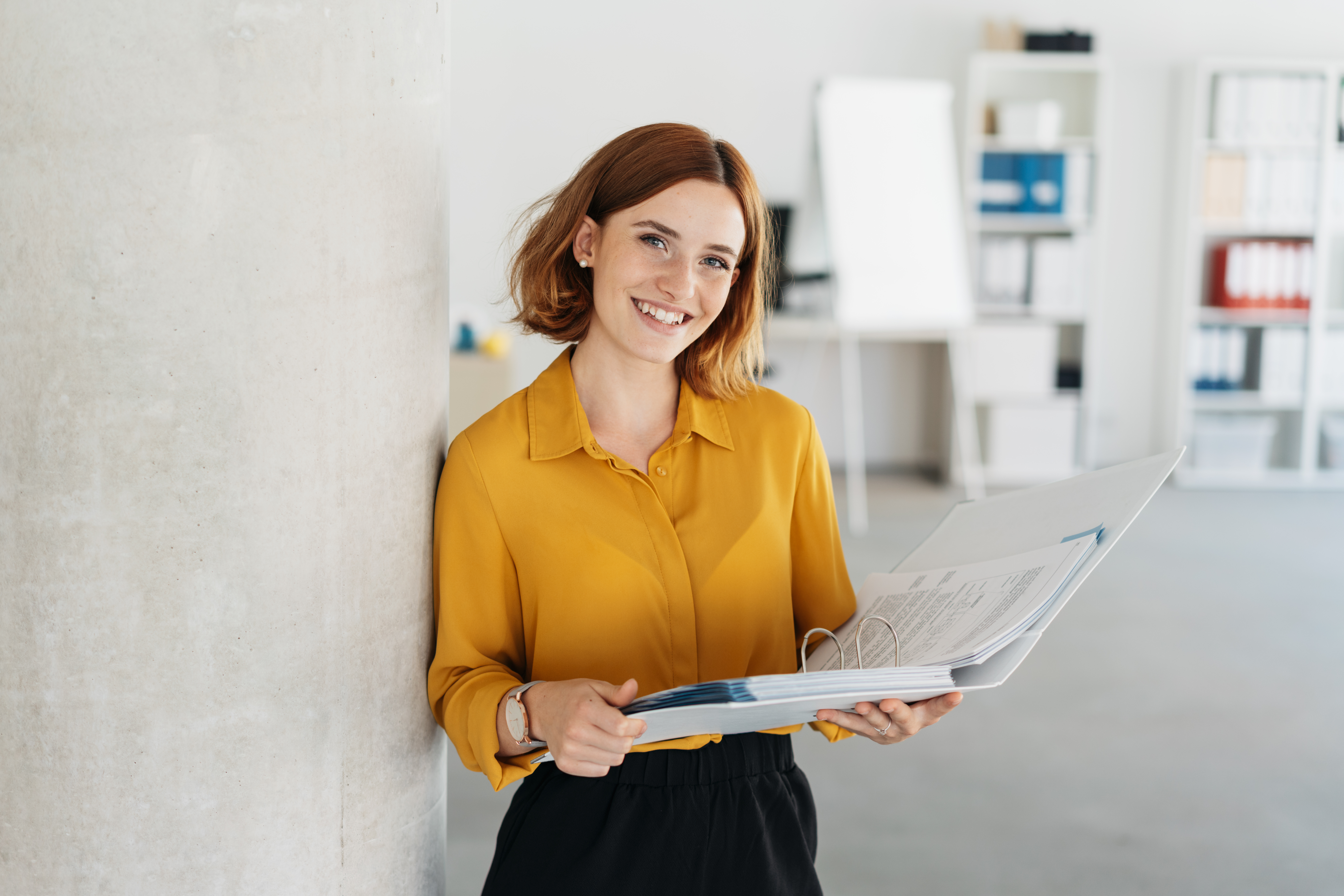 Office worker holding a large open binder as she looks at the camera with a sweet friendly smile